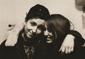 A black and white photo of a young man and woman smiling and embracing, wearing winter clothes, capturing an early Dylan moment.