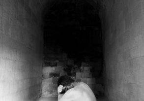 A black and white image of a nude person sitting curled up with their head in their hands at the end of a narrow stone corridor, evoking a sense of emptiness.