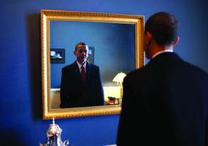 A man in a suit, resembling Barack Obama, looks at his reflection in a large framed mirror in a room with blue walls and a desk lamp.