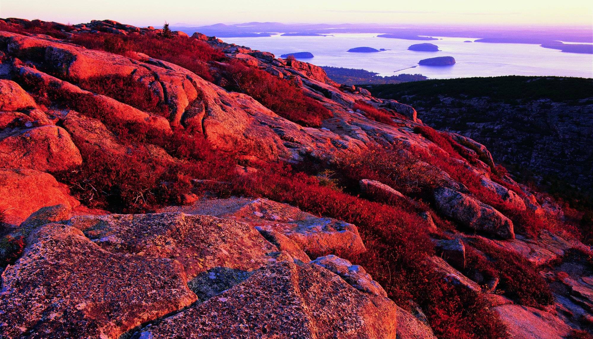 Sunrise illuminates a rocky hillside, highlighting the importance of environmental protection, with a view of distant islands on a calm sea, showcasing rich red and purple hues.