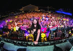 A DJ smiling at a large crowd from behind a West Coast Festival stage setup, with vibrant lights and the audience waving flags and phones in the air.