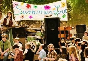 A lively outdoor concert with musicians performing under a "Summer of Love" banner, surrounded by an engaged crowd in a park-like setting.