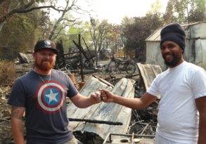 Two men fist bumping in front of a burned-down building in Glen Ellen with charred debris around them. One man wears a Captain America shirt and a cap, while the other wears a white shirt