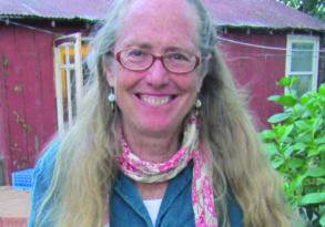 Elderly woman with glasses and long gray hair smiling, wearing a denim shirt and a pink scarf, seated outdoors with a red shed in the background. This is Katrina Frey.