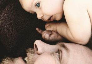 A father's love shining through as he lies on his back with his baby, who is looking into his eyes, resting on his shoulder. Both are against a soft brown background.