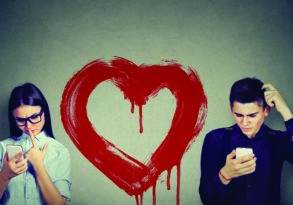 A young man and woman standing back-to-back, looking upset and distracted by their smartphones, embodying a digital disconnect, with a large painted heart dripping between them on a gray wall.