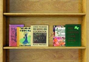 A wooden bookshelf with four colorful cookbooks on two shelves, titled "Joy of Cooking," "Chez Panisse Menu Cookbook," "Ayurvedic Cooking," and "The Greens Cookbook.