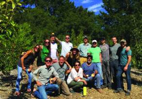 A group of fourteen individuals smiling and standing together in an orchard, holding apples and enjoying a sunny day.