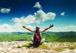 A person wearing a backpack sits on a rocky peak, arms spread wide, facing a vast green landscape under a blue sky with clouds, embodying the spirit of "Connect With Nature.