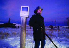 A man in a black coat and hat stands beside a wooden post with a vintage computer monitor on top, in a snowy landscape at dusk in "Cassidy's Tale.