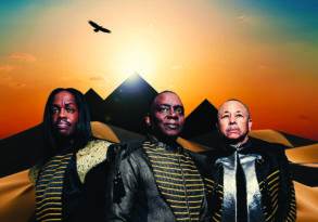 Three men in futuristic armor stand before a desert pyramid landscape at sunset, during the West Coast Festival, with an aircraft flying overhead.