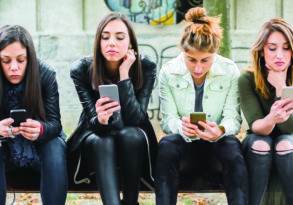 Four women sitting on a bench engrossed in their smartphones, each dressed in casual, modern outfits, likely browsing social media.