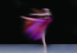 A blurred pictorial of a ballet dancer in motion, wearing a purple and pink dress, captured in a twirl on a dimly lit stage.