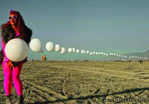 A woman in vibrant pink attire and sunglasses holds a string of large white balloons on a sandy beach with distant mountains.