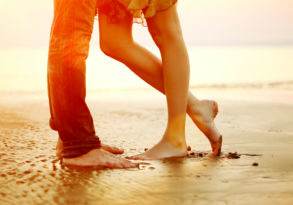 A couple standing barefoot on a beach, legs close up, with a sunset backdrop, in an Ode to Summer.