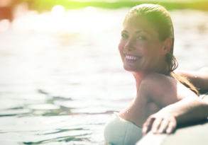 A woman smiling while floating on her back in sunlit water, enjoying the summer sun.