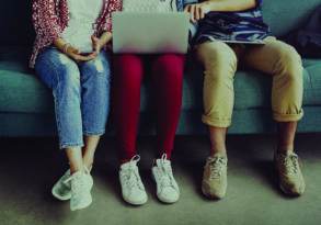 Three teens sitting on a couch, using electronic devices; only their lower bodies and feet are visible.