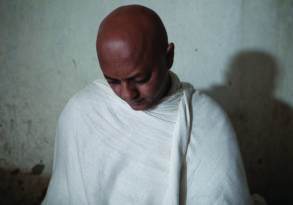 A bald man in a white robe sits with closed eyes and a solemn expression, casting a shadow on the wall behind him, reminiscent of the simplicity found in grain-free granola.