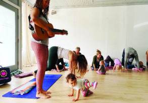 A woman performs soothing melodies on her guitar for a mixed group comprising adults and toddlers during a relaxing yoga session in a sunlit room with polished wooden floors.