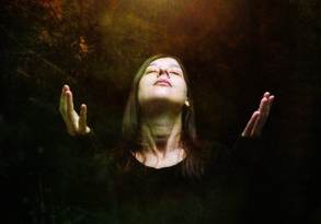 Woman with eyes closed, hands raised, looking upward in a display of faith towards a bright light in a dark, forest-like setting.