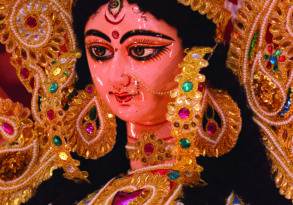 Close-up of a colorful and ornate statue of the Hindu goddess Durga, adorned with intricate jewelry and garments, captured during a Hindu ceremony against a red background.