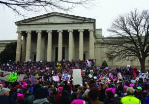 A large crowd of protesters expressing political sentiment, wearing pink hats and holding various signs in front of the national gallery of art during a demonstration.