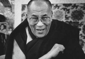 Black and white photo of a smiling elderly man in glasses and traditional robes sitting in front of ornate pearls of consciousness decorations.