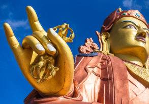 Close-up of a vibrant golden statue’s hand showing a mudra gesture, with the detailed face of the spiritual teacher in the background under a clear blue sky.