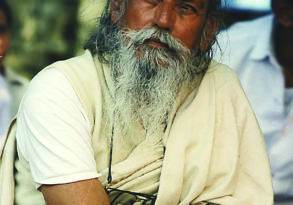 An elderly man, Baba Hari Dass, with a long white beard and mustache, wearing a white robe and red forehead mark, sits with his arms crossed.