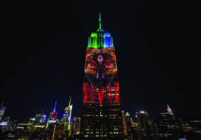 The empire state building at night, illuminated with a colorful light projection of Kali, set against a dark city skyline.