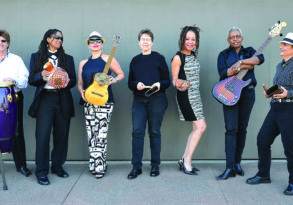 A diverse band of seven musicians with various instruments, including drums, guitars, and a tambourine, posing together against a grey wall adorned with Azúcar con Aché posters.