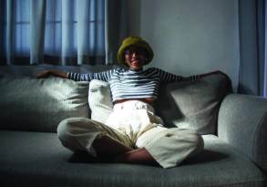 A woman in a striped top, yellow hat, and sunglasses sits smiling on a gray sofa, embodying Black feminist joy, with sunlight filtering through curtains in the background.