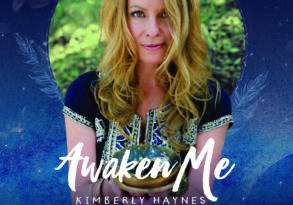 Album cover of "awaken me" by Kimberly Haynes, featuring a portrait of the artist with long blonde hair, smiling in a forest background, framed by feathers and glowing light — epitomizing