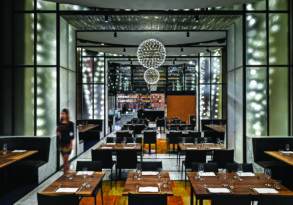 Elegant fine dining restaurant interior featuring geometric furniture and elaborate spherical chandeliers, with soft ambient lighting and a woman walking by.