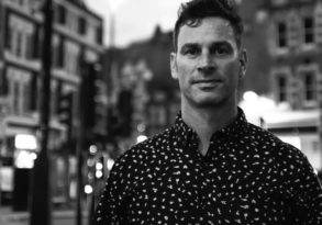 Black and white photo of a man with short hair, speaking wisely and wearing a patterned shirt, standing on a city street at dusk.