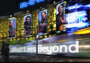 Billboard for R. Carlos Nakai Quartet's album "What Lies Beyond" featuring images of the band members with a cityscape at night in the background, invoking philosophical questions about life or existence