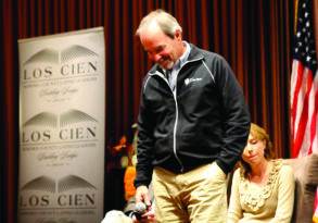 Man smiling as he walks past a seated woman on a stage with a backdrop reading "los cien Sonoma County Latino leaders" and an American flag. Santa Rosa Mayor Chris Coursey is present at