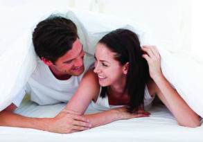 A joyful couple lying under a white sheet, smiling and holding hands, in a brightly lit bedroom after enjoying good sex.