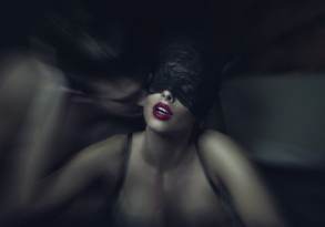 A woman with a kinky lace blindfold and red lipstick appears in a blurred, dynamic scene with a man whispering close to her ear.