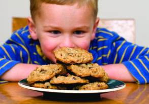 A young boy grinning as he leans towards a stack of chocolate chip cookies on a plate at the dining table, achieving your dreams of indulgence.