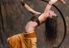 A woman in tribal costume performs an acrobatic pose inside a large metal hoop against a mottled brown backdrop, capturing the wild rhythm of her heritage.