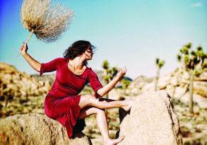 A woman in a red dress leaps between rocks in a desert, holding a large tumbleweed above her head under a clear sky. This scene recalls the iconic setting of the novel "Seductress Re