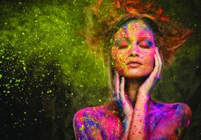 A woman with colorful powder on her face and hair, eyes closed, embodies the artist in everyone against a dark, speckled background.