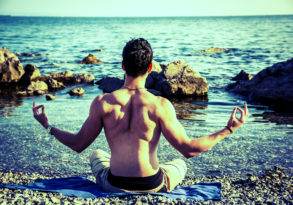 Boy meditating in a lotus pose on a rocky beach facing the sea, with his back towards the camera.