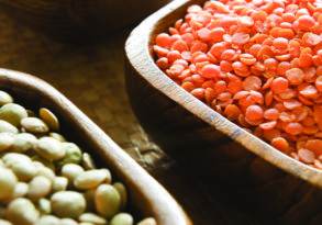 Three wooden bowls containing black beans, red lentils, and mixed legumes, set on a woven mat in an Ayurveda kitchen.