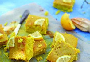 Slices of lemon cornbread from "The Virtuous Tart" on a blue surface, garnished with lemon wedges and garlic cloves, with a knife nearby.