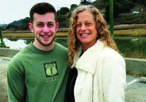 A transgender son and his supportive parent, both smiling, stand together outdoors near a calm water body. The son is in a green shirt, and the parent wears a white jacket.