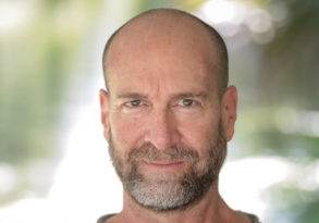 A middle-aged bald man with a beard, giving a subtle smile and staring directly at the camera, with a blurred green background reminiscent of David Deida's serene settings.