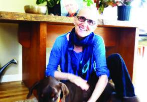 A woman in a blue scarf and boots smiles while sitting beside a large brown dog under a desk with potted plants, reminiscing about her sessions as a Tantra Teacher.