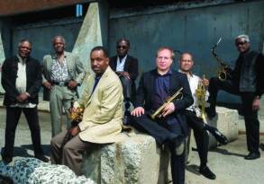 Seven middle-aged men, including a trumpet player and two saxophonists from The Cookers, posing together outdoors, dressed in semi-formal attire.